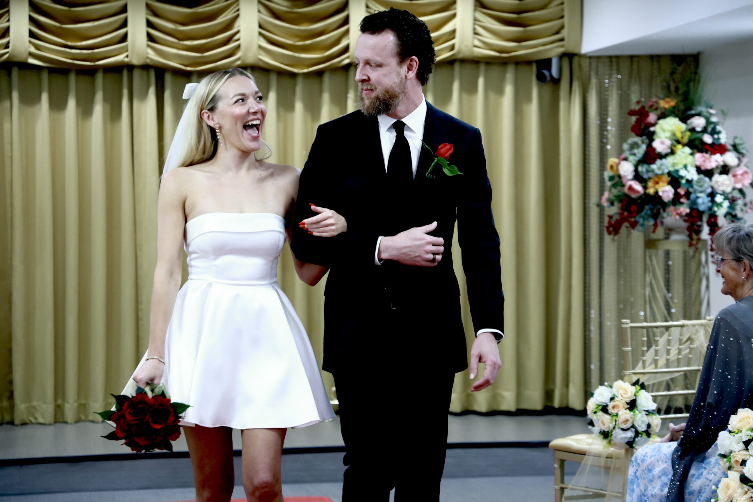 Bride in a white dress smiles joyfully while walking arm-in-arm with a groom in a tuxedo. A guest watches from the side, adding to the celebratory atmosphere.