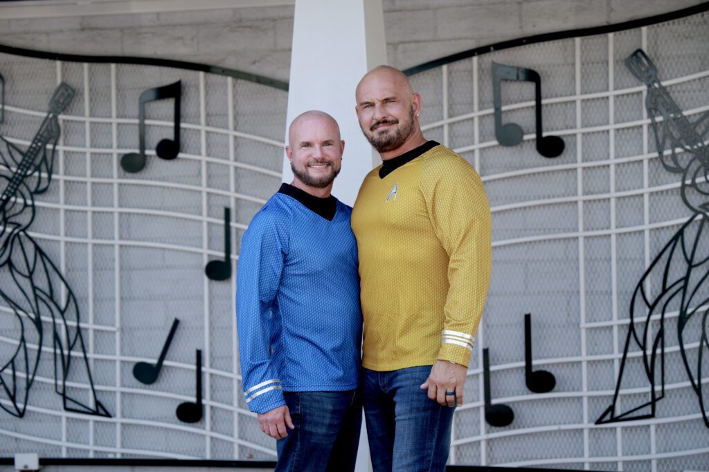 Two men pose together in vibrant blue and gold Starfleet uniforms against a backdrop of musical notes, highlighting a fun, themed event.