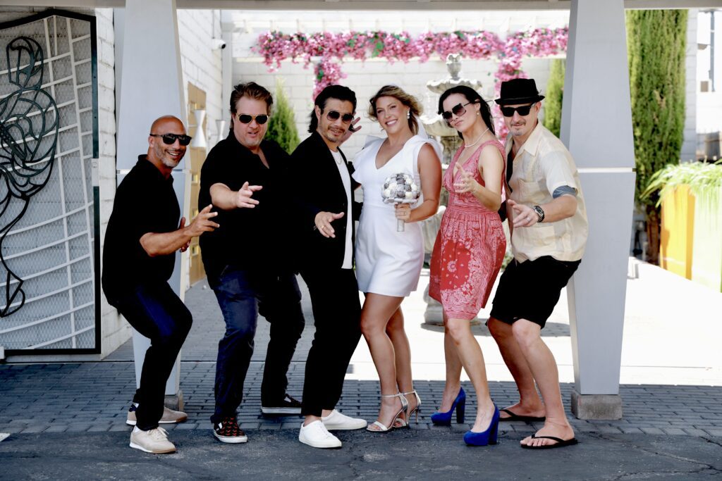 A joyful group of six friends poses together, dressed stylishly for a celebration, with a bride in a white dress holding a bouquet.