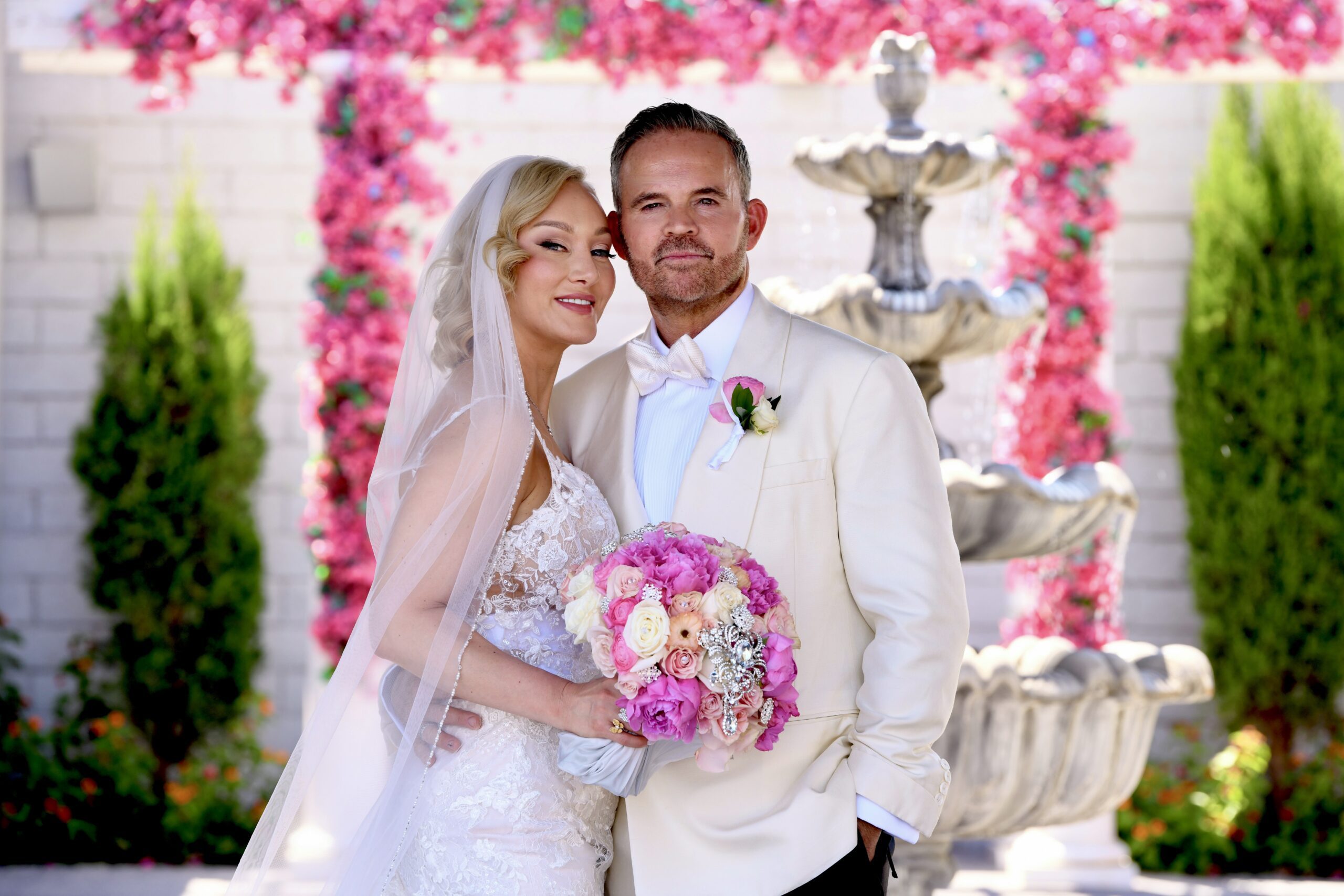 Bride and groom pose together in a picturesque outdoor setting, adorned with pink flowers and a fountain, celebrating their wedding day.