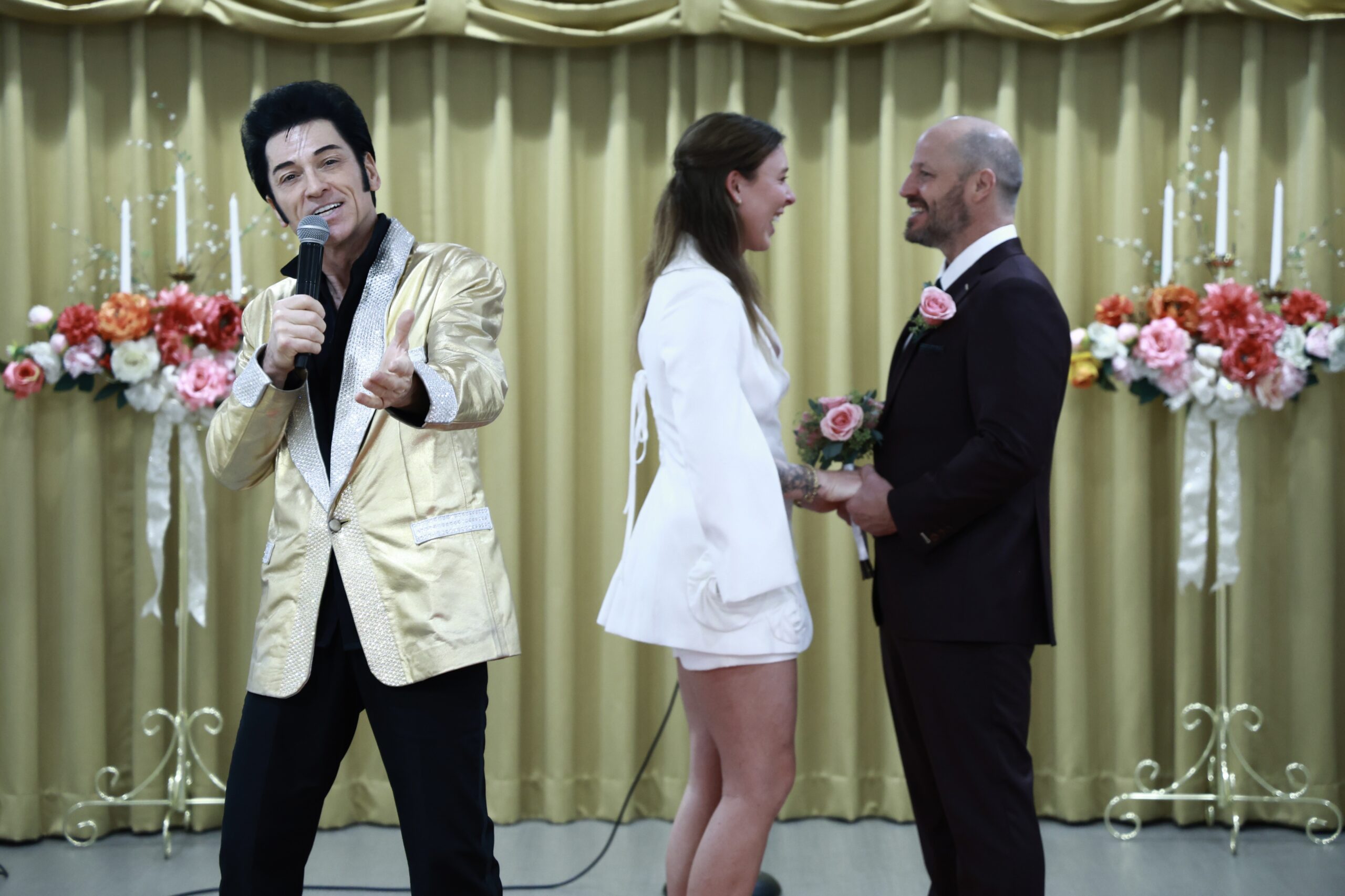 An Elvis impersonator sings during a wedding ceremony, with a bride in a white dress and a groom holding flowers, set against a floral backdrop.