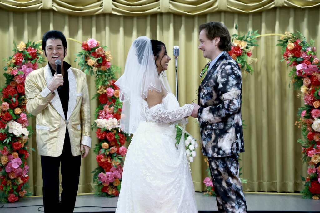A bride in a white lace gown smiles at her groom, who wears a patterned suit, during a wedding ceremony officiated by a man in a shiny gold jacket. Floral decorations frame the scene.
