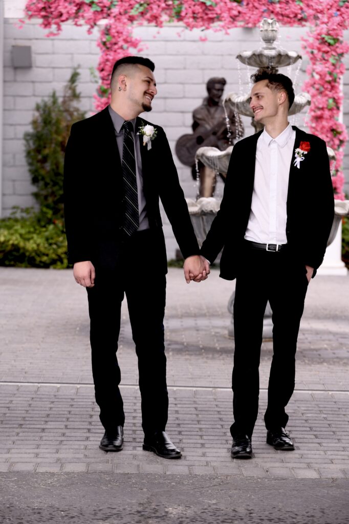 Two smiling men in formal attire hold hands, standing by a decorative fountain, surrounded by pink flowers, symbolizing love and celebration.