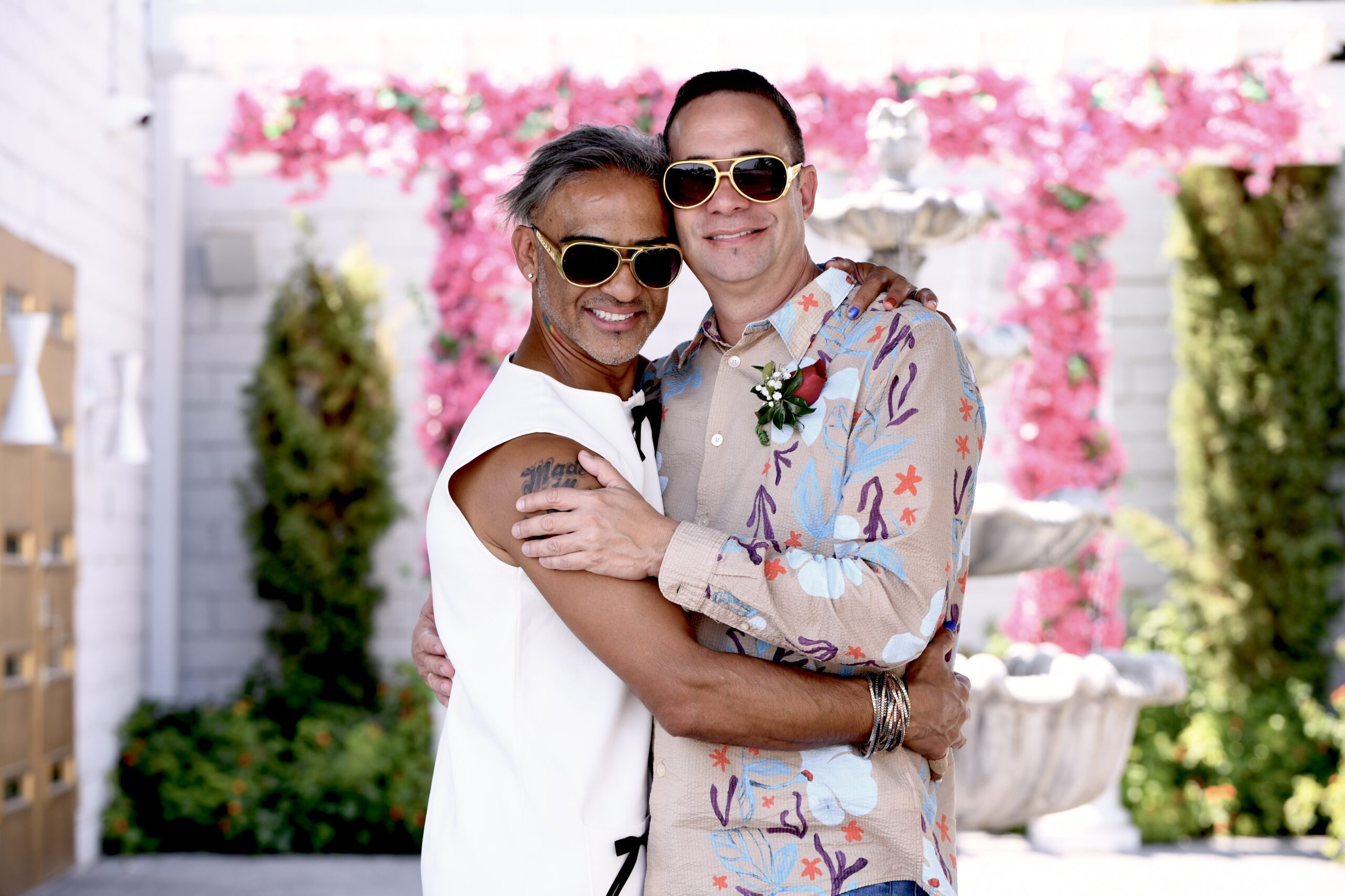 Two men in sunglasses embrace joyfully in front of Elvis Chapel fountain adorned with pink flowers.