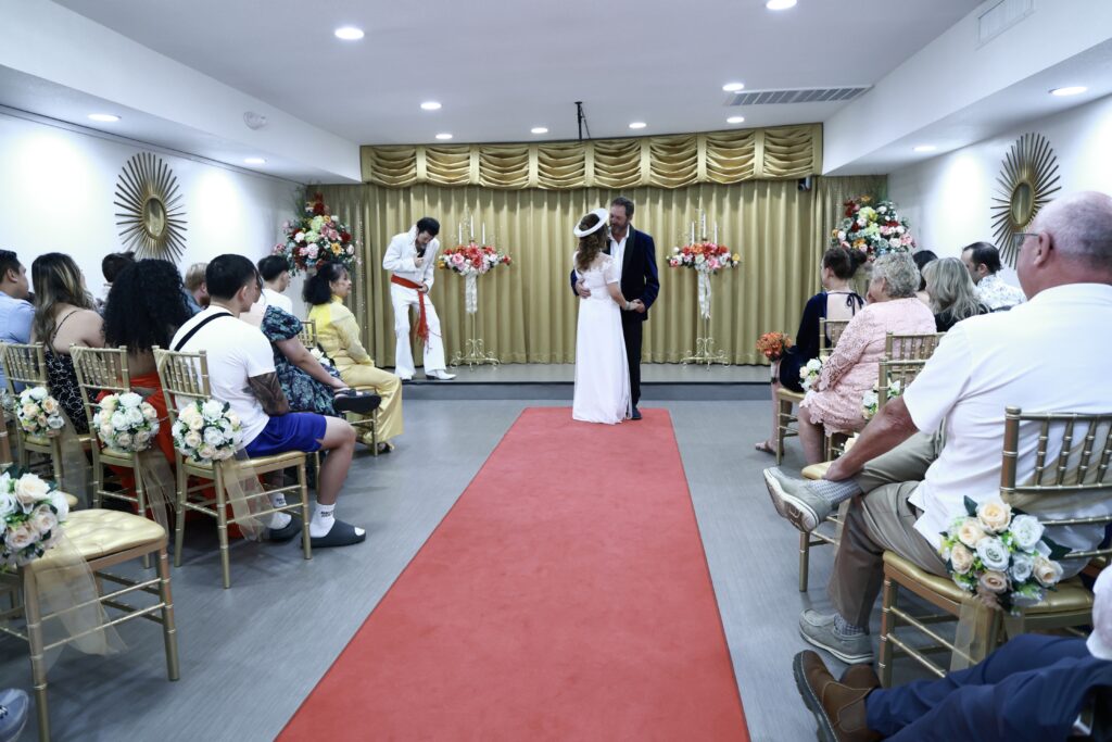 A couple exchanges vows on a red carpet during their Elvis wedding ceremony in Las Vegas chapel, attended by guests. An officiant in white leads the event at a festive venue.