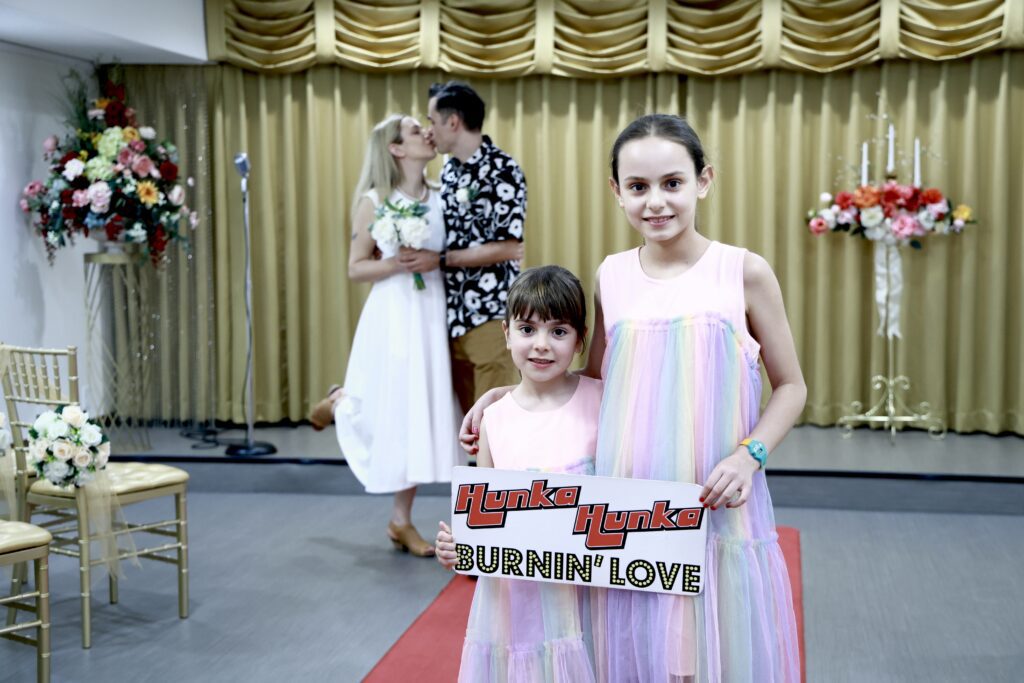 A bride and groom kiss in the background of Elvis wedding ceremony in Las Vegas chapel, while two girls in pastel dresses hold a sign reading "Hunka Hunka Burnin' Love," celebrating the wedding.