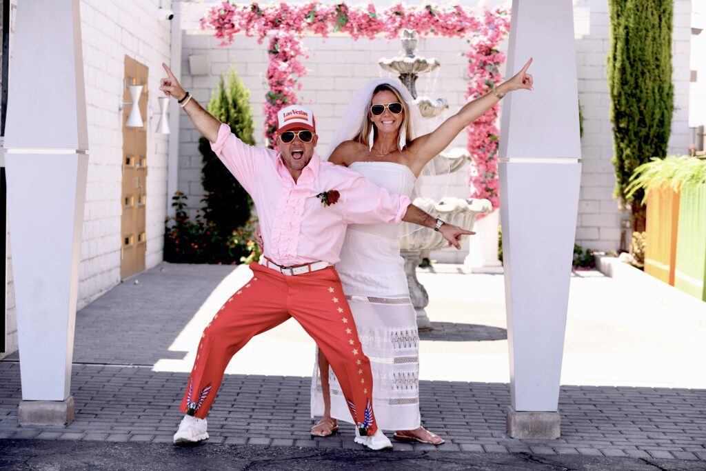 A man in a pink ruffled shirt and bright red pants poses playfully with a woman in a white dress and veil, celebrating a fun wedding moment at Elvis wedding ceremony in Las Vegas chapel.