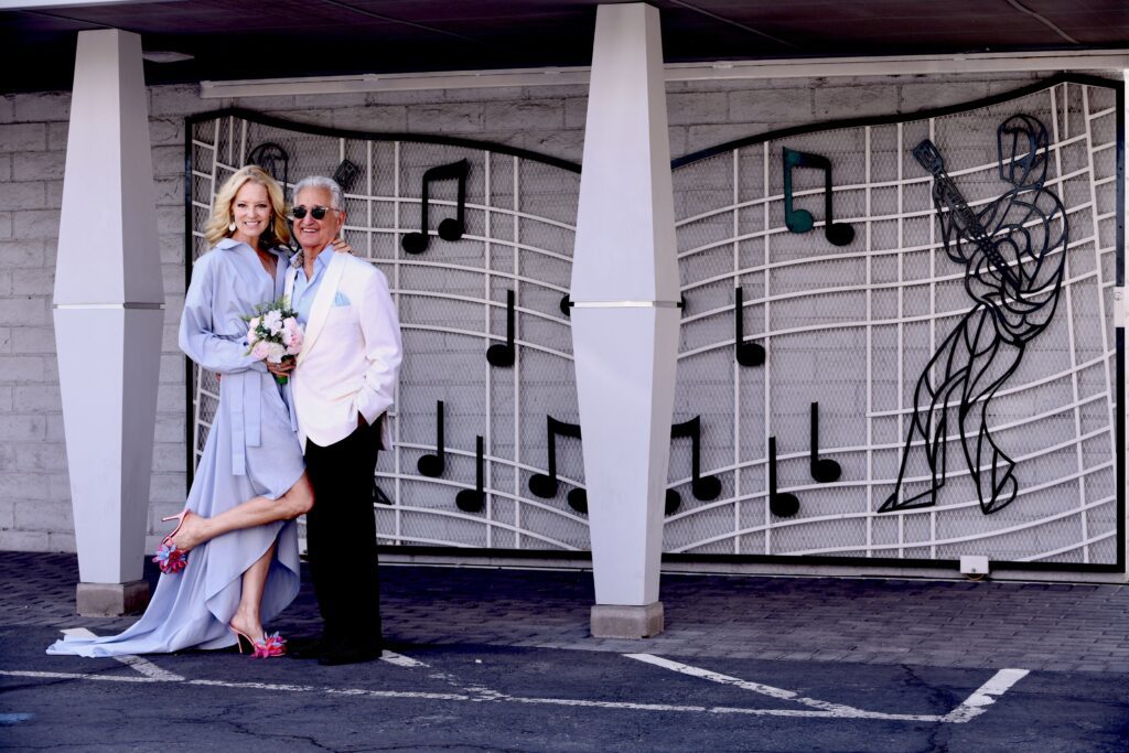 A smiling couple poses in front of a musical-themed backdrop Elvis wedding ceremony, showcasing a decorative metal design of a musician and musical notes.