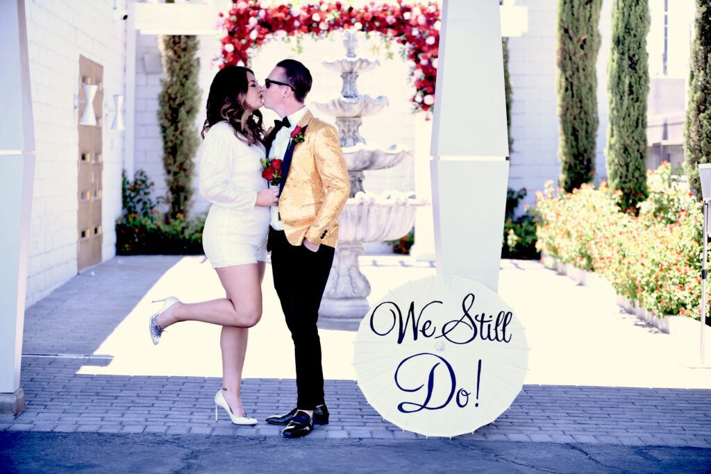 A couple shares a kiss in a romantic Elvis wedding ceremony outdoor setting, celebrating their love with a sign that reads "We Still Do!" amidst floral decorations.